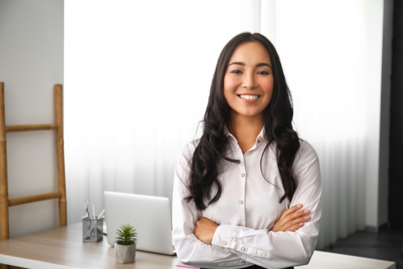 "Professional woman in a bright office with arms crossed, smiling confidently in front of a laptop, representing content marketing services.