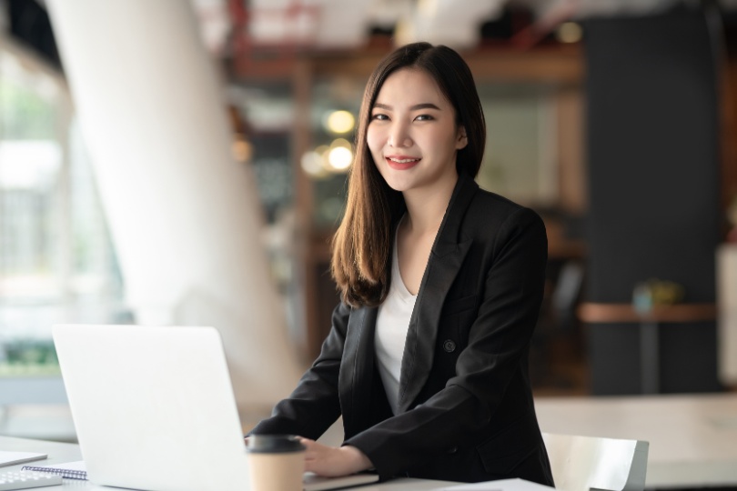 A professional woman sitting at a desk with a laptop and coffee cup, smiling confidently in a modern office setting, representing Social Media Marketing Services.